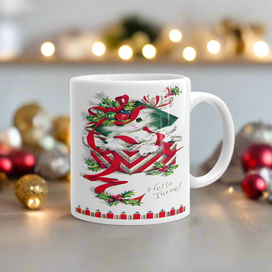 This ceramic glossy coffee mug features a midcentry retro Christmas illustration that says Hello There! with a white kitten sitting in a red and white striped giftbox with a green lid on her head.