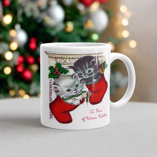 This white ceramic glossy coffee mug features two adorable kittens in red stockings at a Christmas mantle. The print is retro kitsch and says A Pair of Warm Wishes.