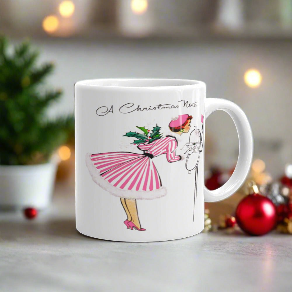 This ceramic glossy coffee mug features a retro vintage Christmas illustration of a woman holding a holly plant checking her mailbox. She is wearing a pink hat, pink and white striped dress, and pink heels. The illustration says A Christmas Note.