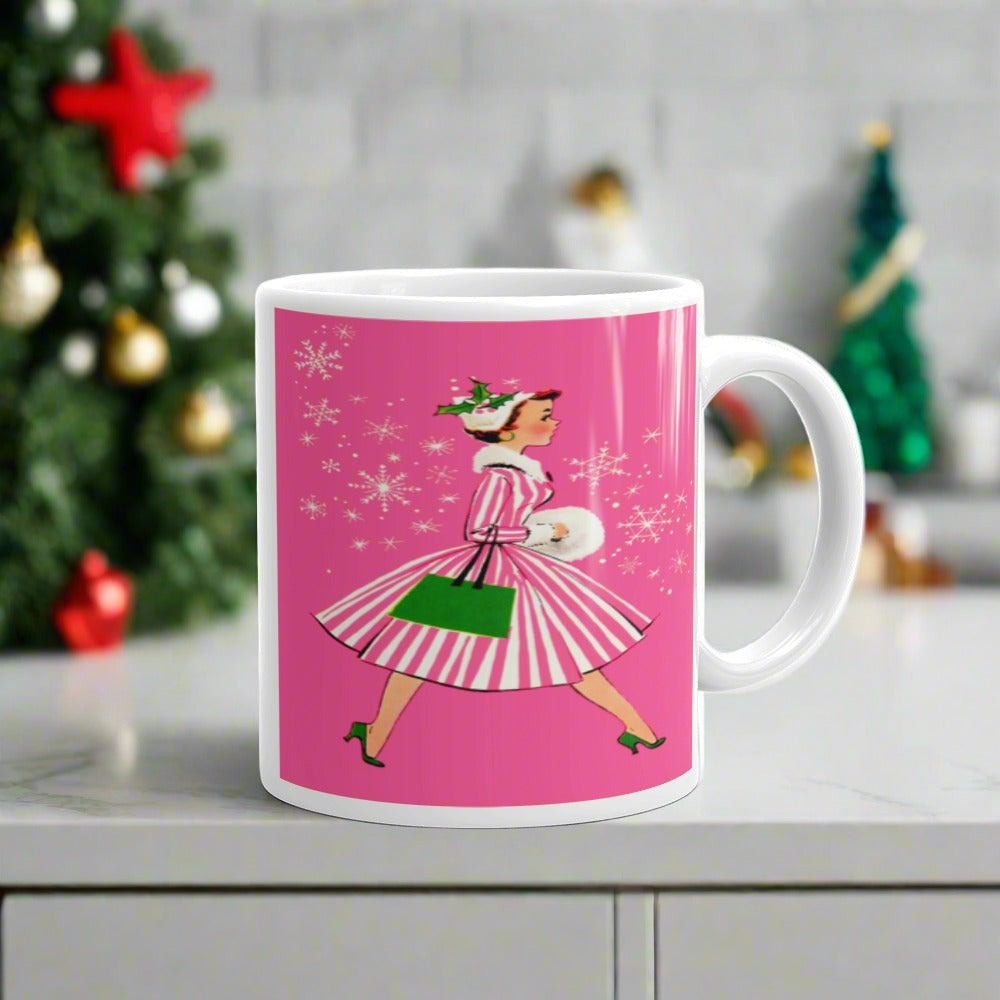 This ceramic glossy mug features a vintage Christmas illustration of a retro Midcentury housewife dressed in a pink and white striped dress with a green purse and heels. The background is pink and there are snowflakes. 