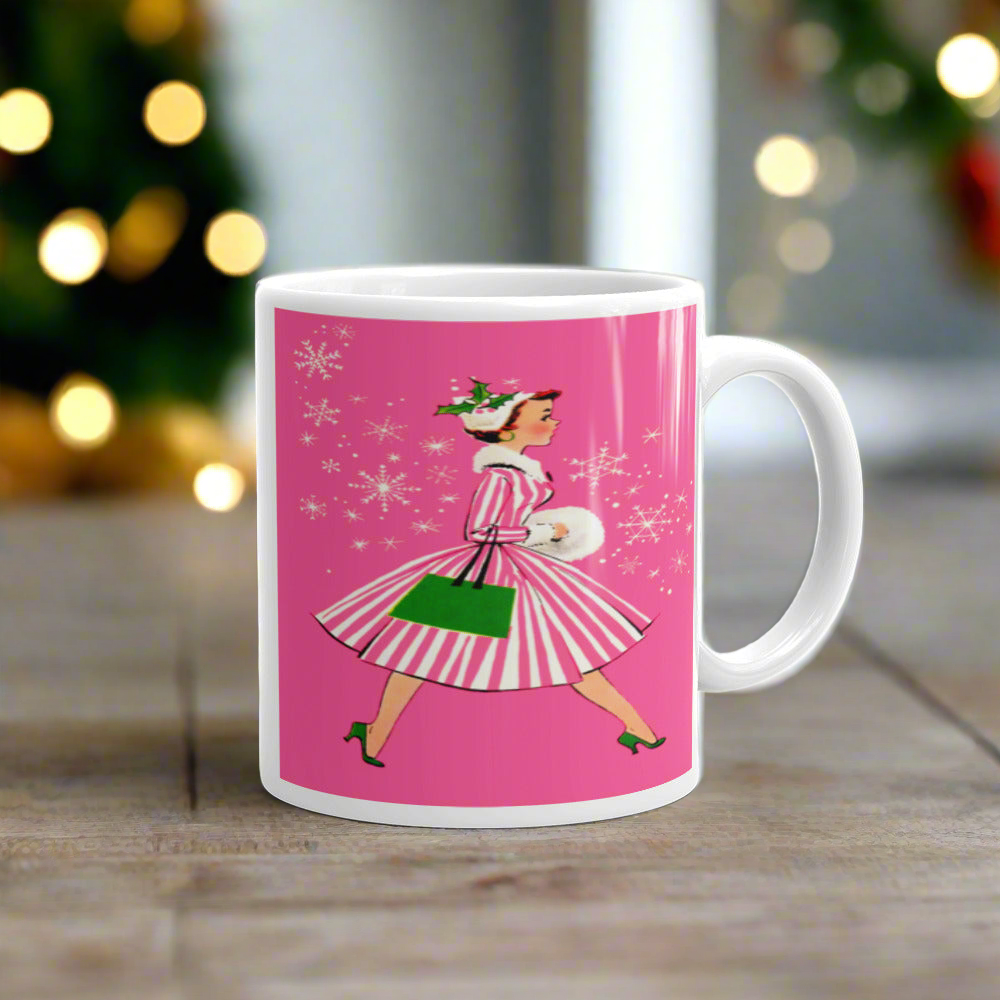 This ceramic glossy mug features a vintage Christmas illustration of a retro Midcentury housewife dressed in a pink and white striped dress with a green purse and heels. The background is pink and there are snowflakes. 