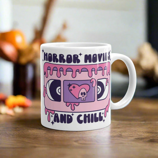 This ceramic glossy coffee mug features a Halloween print that captures a retro 70s feel. There is a pink and purple VHS tape with a heart and skeleton in the center and around the tape it says Horror Movies and Chill. The tape is dripping with pink blood.