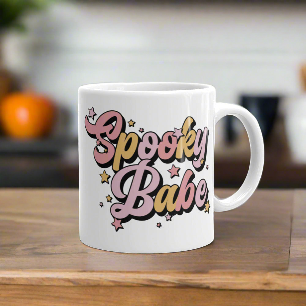 This ceramic glossy coffee mug features cursive lettering that says Spooky Babe. The colors are pastel pinks and yellows and have stars.