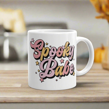 This ceramic glossy coffee mug features cursive lettering that says Spooky Babe. The colors are pastel pinks and yellows and have stars.