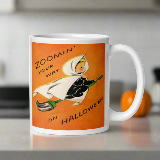 This ceramic glossy coffee mug features a vintage illustration of an adorable little blond witch in a white cape flying on a broom. The background is orange and says Zoomin' your way on Halloween.