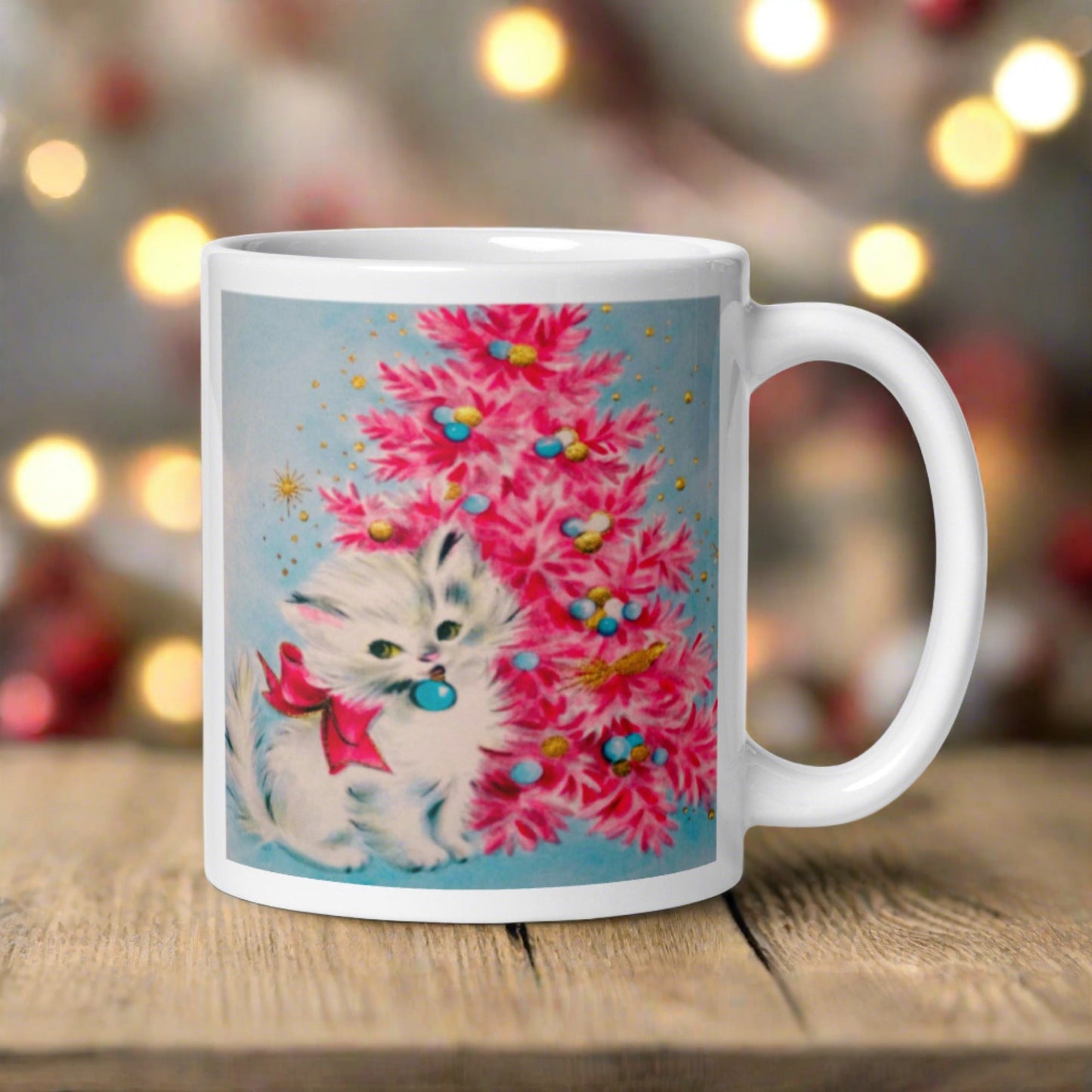 This glossy ceramic coffee cup features an adorable vintage retro print of a white christmas kitten with a blue ornament in her mouth. She is wearing a pink bow and is sitting next to a pink Christmas tree with a blue background.