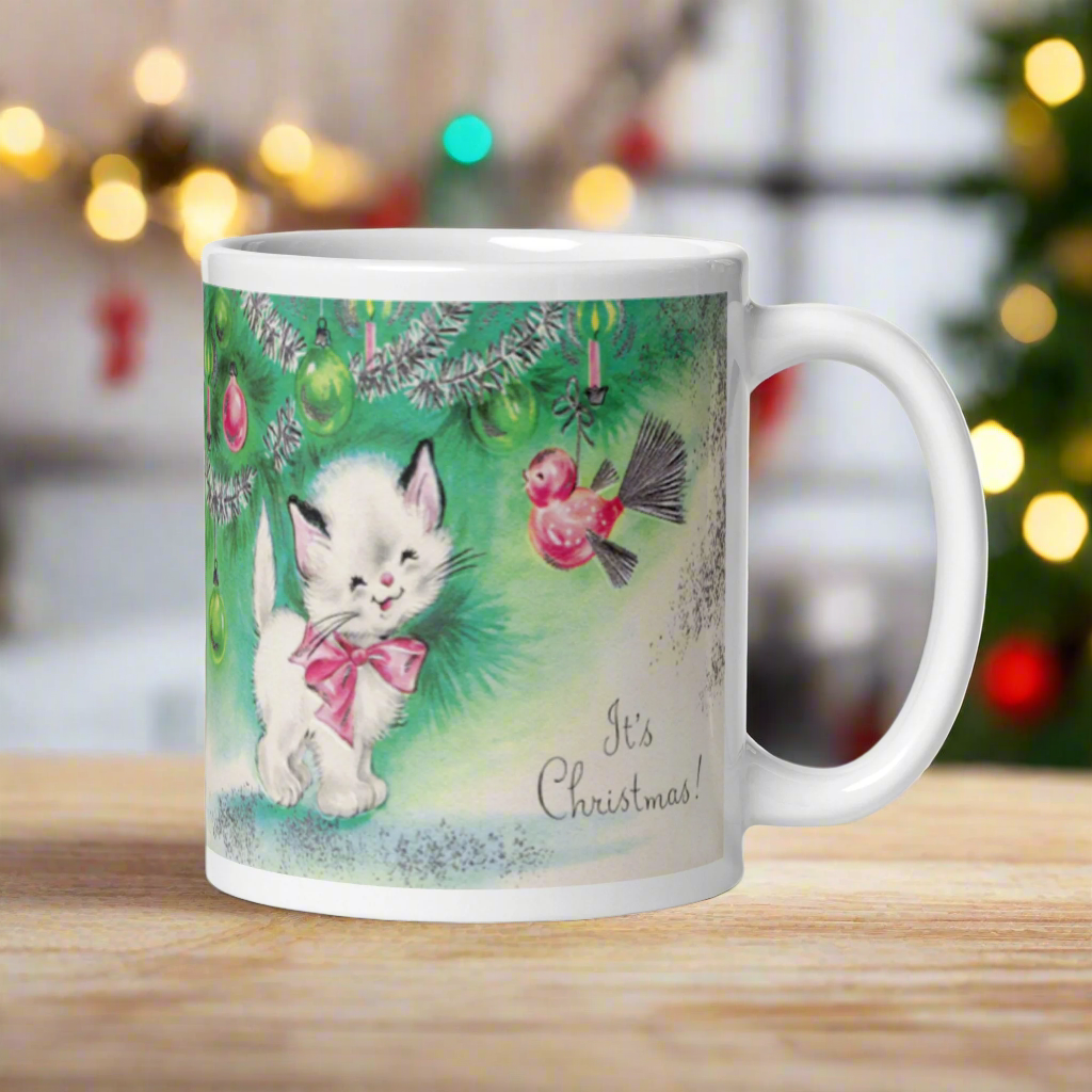 This ceramic glossy coffee mug features a midcentury vintage Christmas Illustration that says It's Christmas! There is an adorable white kitten wearing a pink bow underneath the christmas tree.