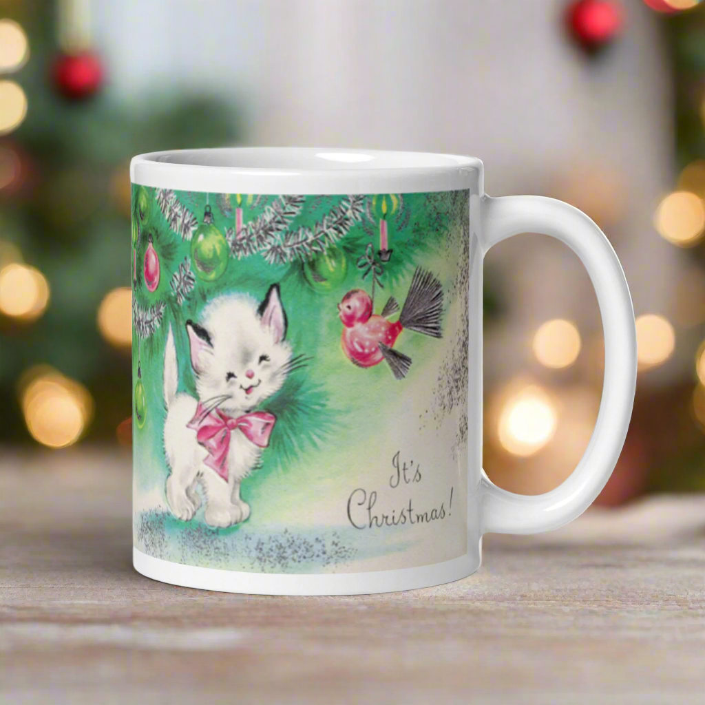 This ceramic glossy coffee mug features a midcentury vintage Christmas Illustration that says It's Christmas! There is an adorable white kitten wearing a pink bow underneath the christmas tree.