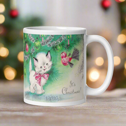 This ceramic glossy coffee mug features a midcentury vintage Christmas Illustration that says It's Christmas! There is an adorable white kitten wearing a pink bow underneath the christmas tree.