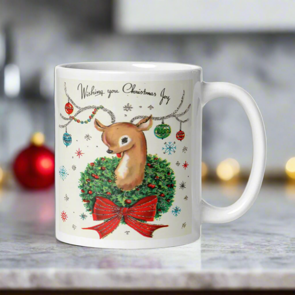 This ceramic glossy coffee mug is a midcentury christmas illustration that says Wishing you Christmas Joy. It features a reindeer with her tongue out wearing a Christmas wreath with a red bow. Her antlers are decorated with tinsel and  ornaments.