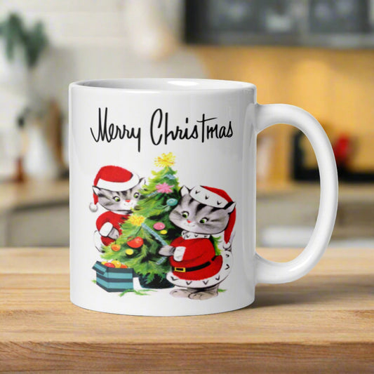 This glossy ceramic coffee mug features a vintage illustration of two cats in santa outfits decorating their Christmas tree. It says Merry Christmas above the artwork in a Midcentury style lettering.