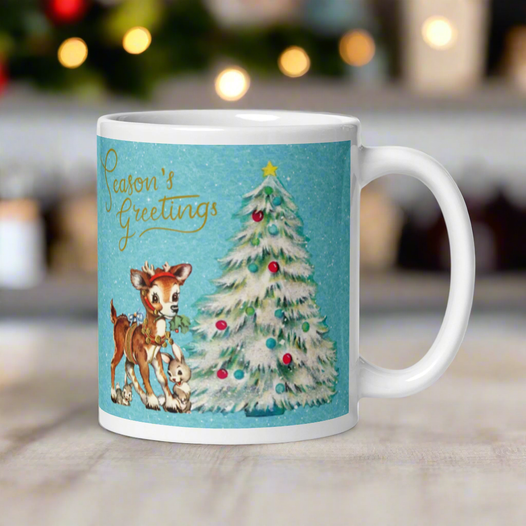 This glossy ceramic coffee mug features 
 Midcentury vintage artwork of an adorable reindeer with holly in her mouth, bunnies, and a Christmas Tree. There is gold lettering that says Season's Greetings and a blue felt background.