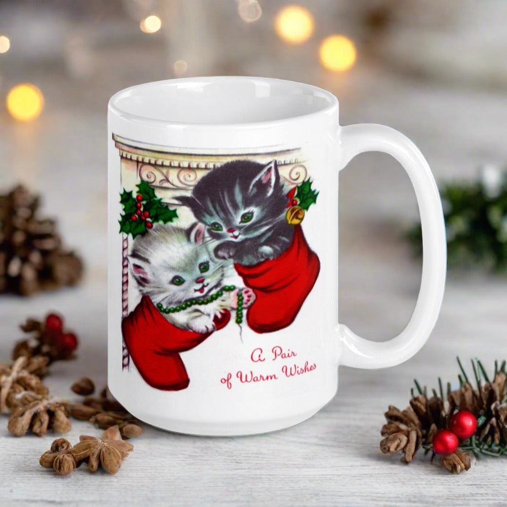 This white ceramic glossy coffee mug features two adorable kittens in red stockings at a Christmas mantle. The print is retro kitsch and says A Pair of Warm Wishes.