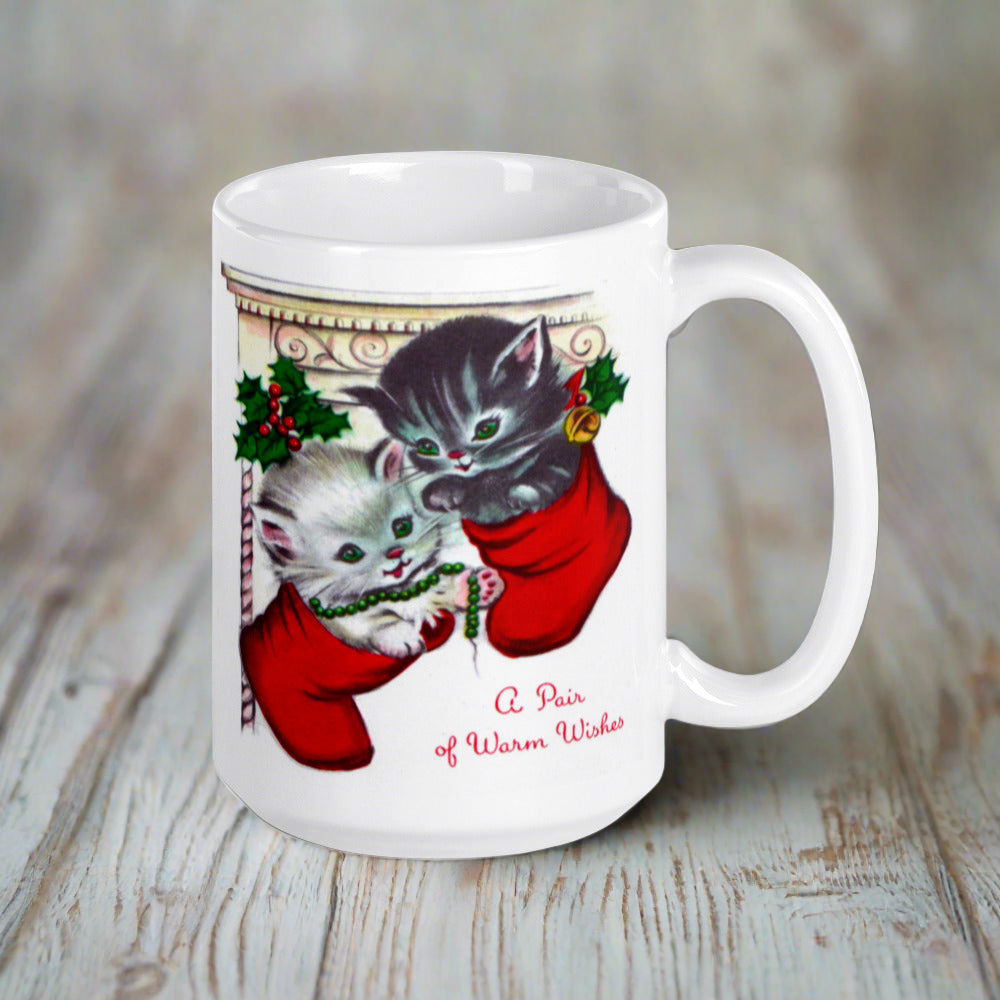 This white ceramic glossy coffee mug features two adorable kittens in red stockings at a Christmas mantle. The print is retro kitsch and says A Pair of Warm Wishes.