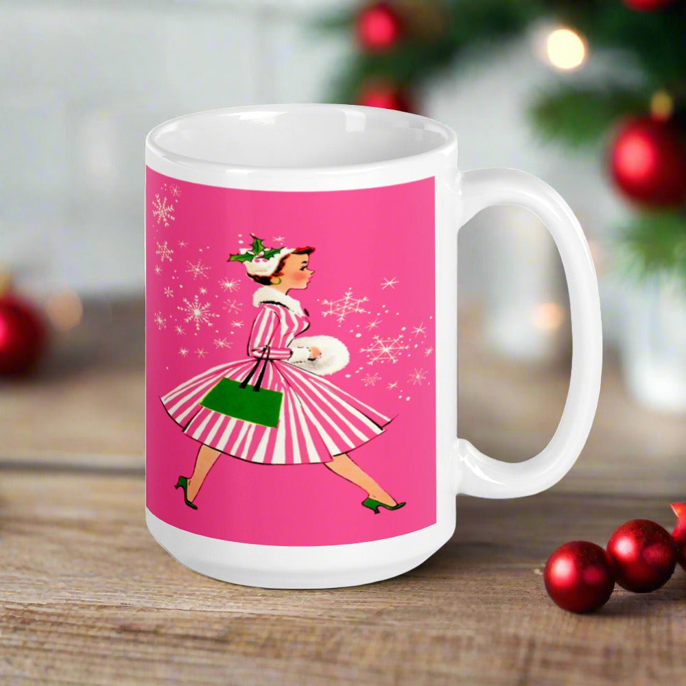 This ceramic glossy mug features a vintage Christmas illustration of a retro Midcentury housewife dressed in a pink and white striped dress with a green purse and heels. The background is pink and there are snowflakes. 