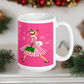 This ceramic glossy mug features a vintage Christmas illustration of a retro Midcentury housewife dressed in a pink and white striped dress with a green purse and heels. The background is pink and there are snowflakes. 