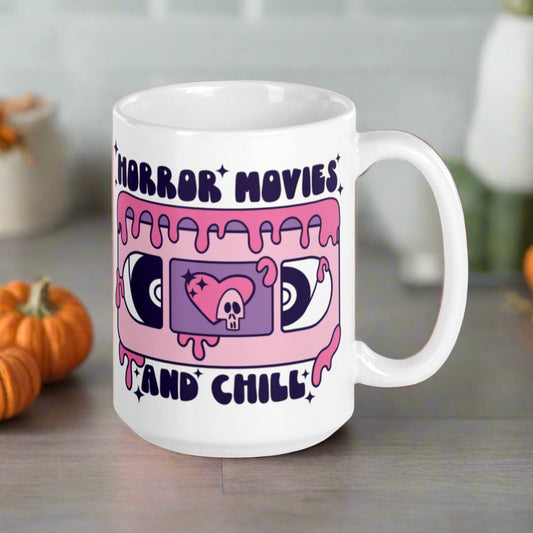 This ceramic glossy coffee mug features a Halloween print that captures a retro 70s feel. There is a pink and purple VHS tape with a heart and skeleton in the center and around the tape it says Horror Movies and Chill. The tape is dripping with pink blood.