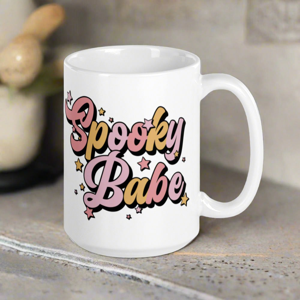 This ceramic glossy coffee mug features cursive lettering that says Spooky Babe. The colors are pastel pinks and yellows and have stars.