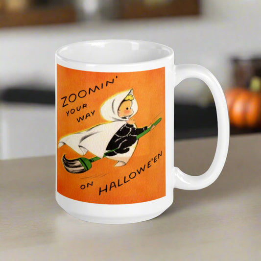 This ceramic glossy coffee mug features a vintage illustration of an adorable little blond witch in a white cape flying on a broom. The background is orange and says Zoomin' your way on Halloween.