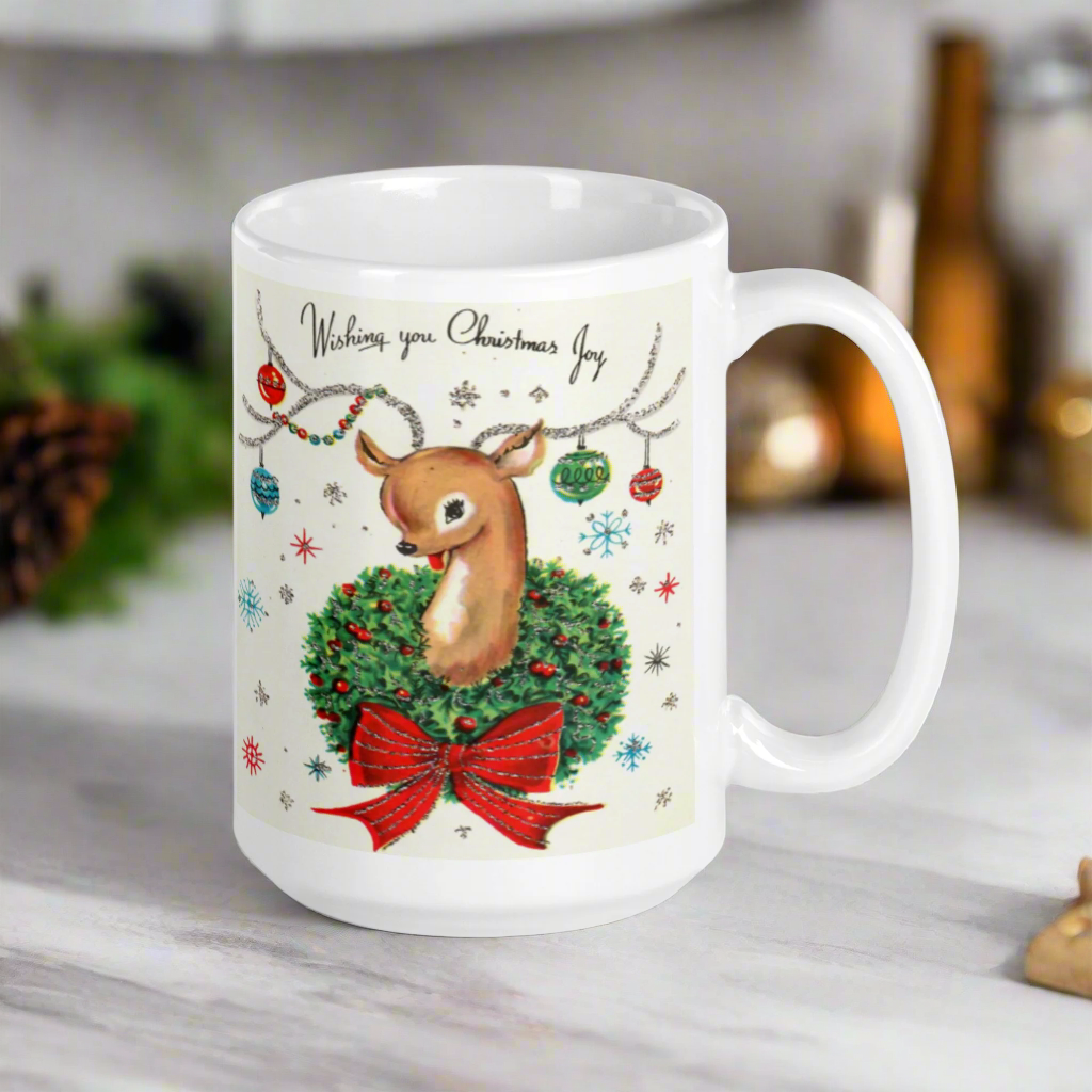 This ceramic glossy coffee mug is a midcentury christmas illustration that says Wishing you Christmas Joy. It features a reindeer with her tongue out wearing a Christmas wreath with a red bow. Her antlers are decorated with tinsel and  ornaments.