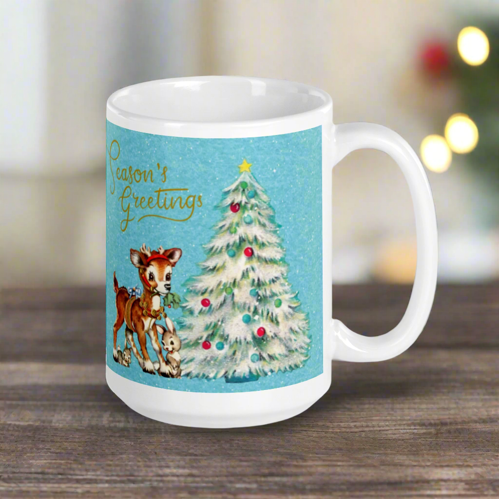 This glossy ceramic coffee mug features 
 Midcentury vintage artwork of an adorable reindeer with holly in her mouth, bunnies, and a Christmas Tree. There is gold lettering that says Season's Greetings and a blue felt background.