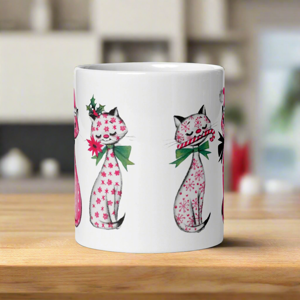 This ceramic glossy coffee mug features a midcentury retro Christmas illustration of three cats. They are adorned with bows around their necks. One has a pink santa hat on her head, one has a candy cane in her mouth and the other is holding a flower.