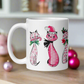 This ceramic glossy coffee mug features a midcentury retro Christmas illustration of three cats. They are adorned with bows around their necks. One has a pink santa hat on her head, one has a candy cane in her mouth and the other is holding a flower.