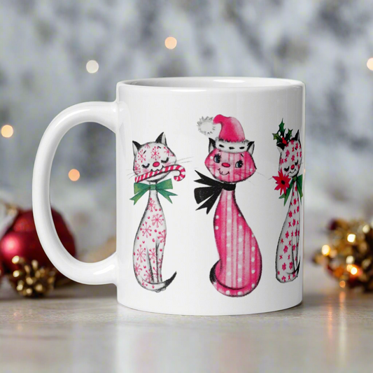 This ceramic glossy coffee mug features a midcentury retro Christmas illustration of three cats. They are adorned with bows around their necks. One has a pink santa hat on her head, one has a candy cane in her mouth and the other is holding a flower.