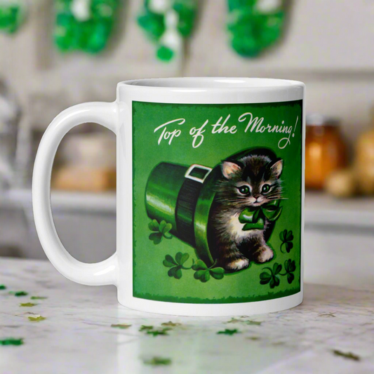 This ceramic glossy coffee mug features a midcentury retro St. Patrick's day illustration that says Top of the Morning! with a kitten sitting in a leprechaun hat surrounded by shamrocks.