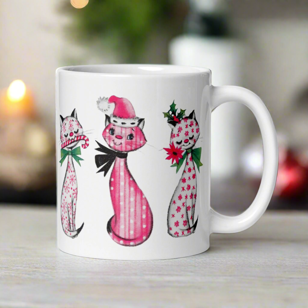 This ceramic glossy coffee mug features a midcentury retro Christmas illustration of three cats. They are adorned with bows around their necks. One has a pink santa hat on her head, one has a candy cane in her mouth and the other is holding a flower.