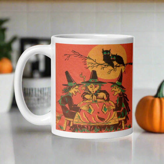 This ceramic glossy mug features a vintage Halloween print of three witches sitting around a large jack o'lantern. There are two owls sitting in a tree branch above them with a full yellow moon.