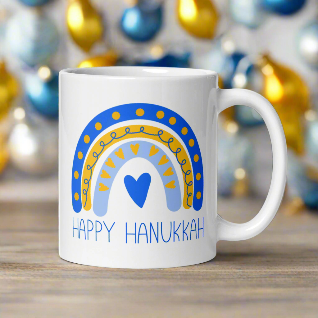 This ceramic glossy coffee cup says Happy Hanukkah and features a blue and yellow boho rainbow with a heart.