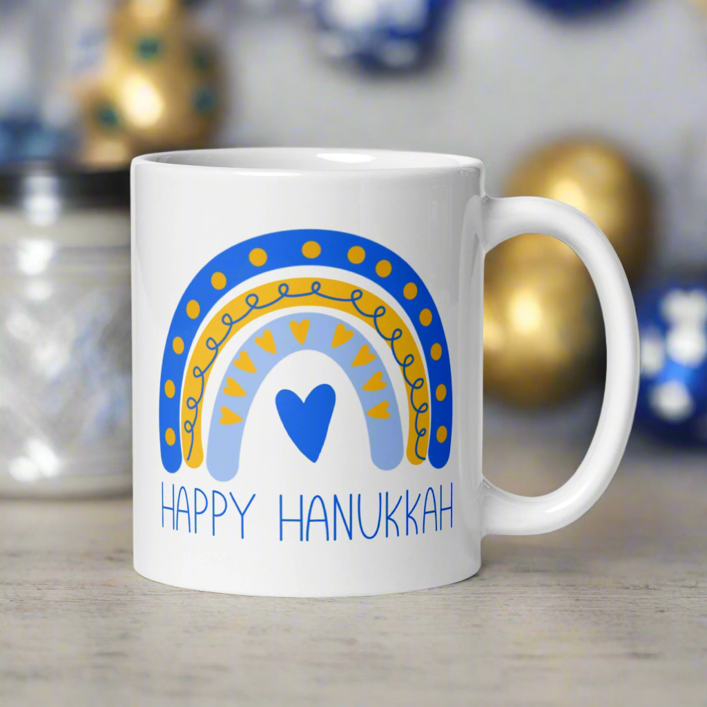 This ceramic glossy coffee cup says Happy Hanukkah and features a blue and yellow boho rainbow with a heart.