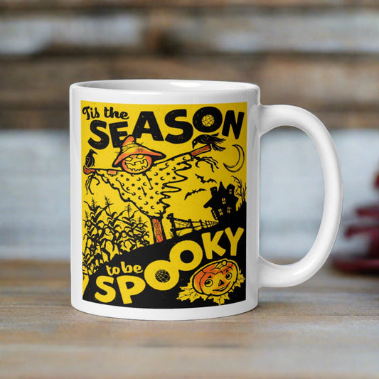 This ceramic glossy coffee mug features a retro vintage print that says Tis the Season to be Spooky. It is yellow and black and features a scarecrow, a cute jack o lantern and a spooky house in the background.