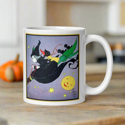 This ceramic glossy mug features a vintage Halloween illustration by Fern Bisel Peat. It features a witch dressed in all black soaring through the air on her broom with black cats. There is a smiling full yellow moon.