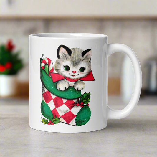 This ceramic glossy coffee mug features a retro vintage Midcentury illustration of an adorable kitten wearing a red bow and is in a stocking with a candy cane.