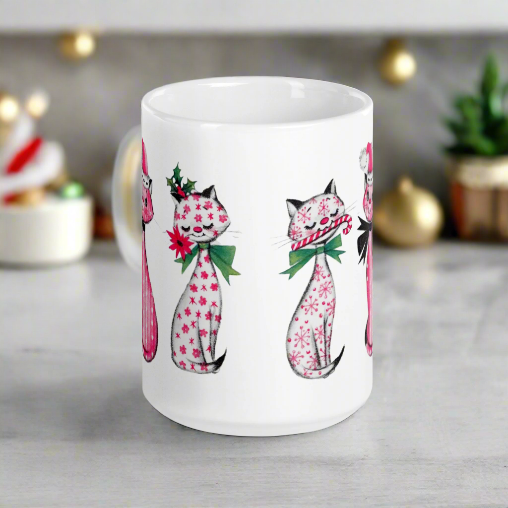 This ceramic glossy coffee mug features a midcentury retro Christmas illustration of three cats. They are adorned with bows around their necks. One has a pink santa hat on her head, one has a candy cane in her mouth and the other is holding a flower.