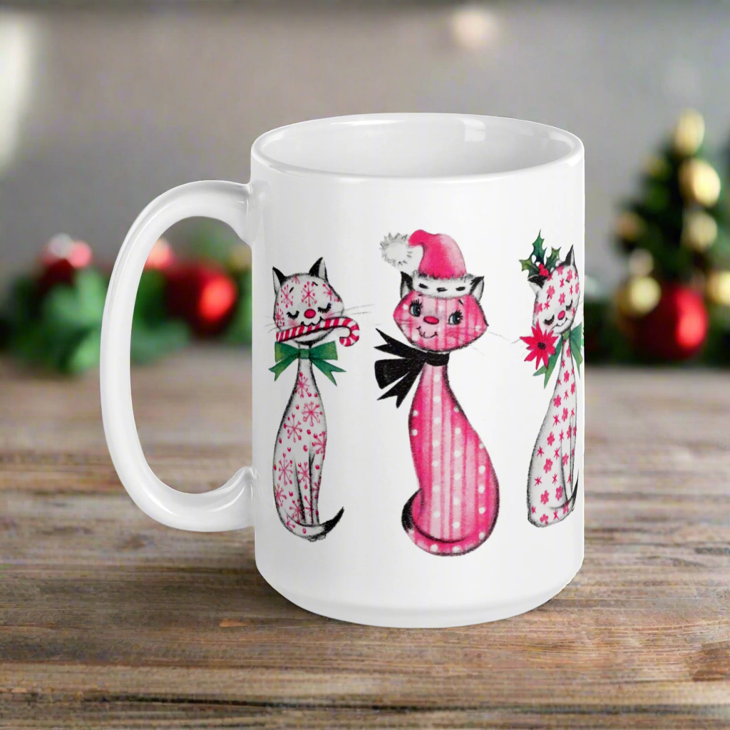 This ceramic glossy coffee mug features a midcentury retro Christmas illustration of three cats. They are adorned with bows around their necks. One has a pink santa hat on her head, one has a candy cane in her mouth and the other is holding a flower.