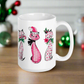 This ceramic glossy coffee mug features a midcentury retro Christmas illustration of three cats. They are adorned with bows around their necks. One has a pink santa hat on her head, one has a candy cane in her mouth and the other is holding a flower.