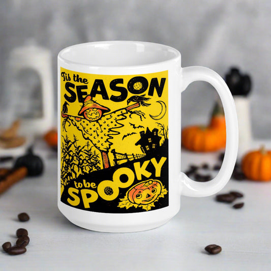 This ceramic glossy coffee mug features a retro vintage print that says Tis the Season to be Spooky. It is yellow and black and features a scarecrow, a cute jack o lantern and a spooky house in the background.