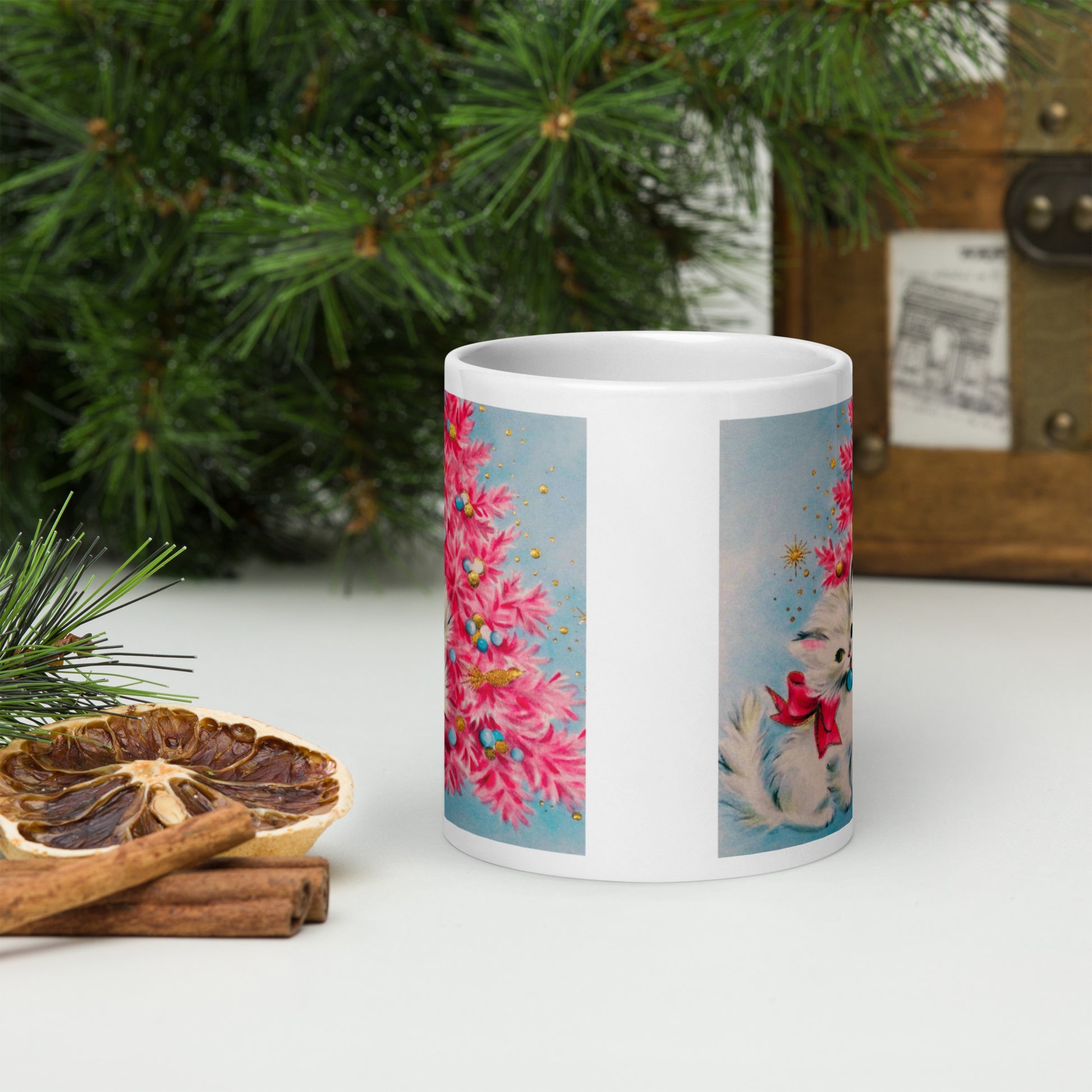 This glossy ceramic coffee cup features an adorable vintage retro print of a white christmas kitten with a blue ornament in her mouth. She is wearing a pink bow and is sitting next to a pink Christmas tree with a blue background.