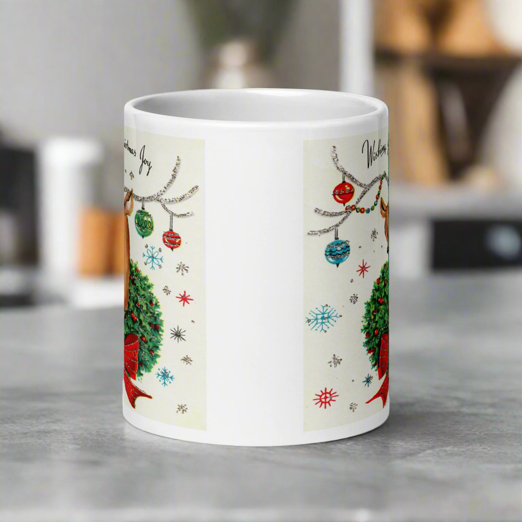 This ceramic glossy coffee mug is a midcentury christmas illustration that says Wishing you Christmas Joy. It features a reindeer with her tongue out wearing a Christmas wreath with a red bow. Her antlers are decorated with tinsel and  ornaments.