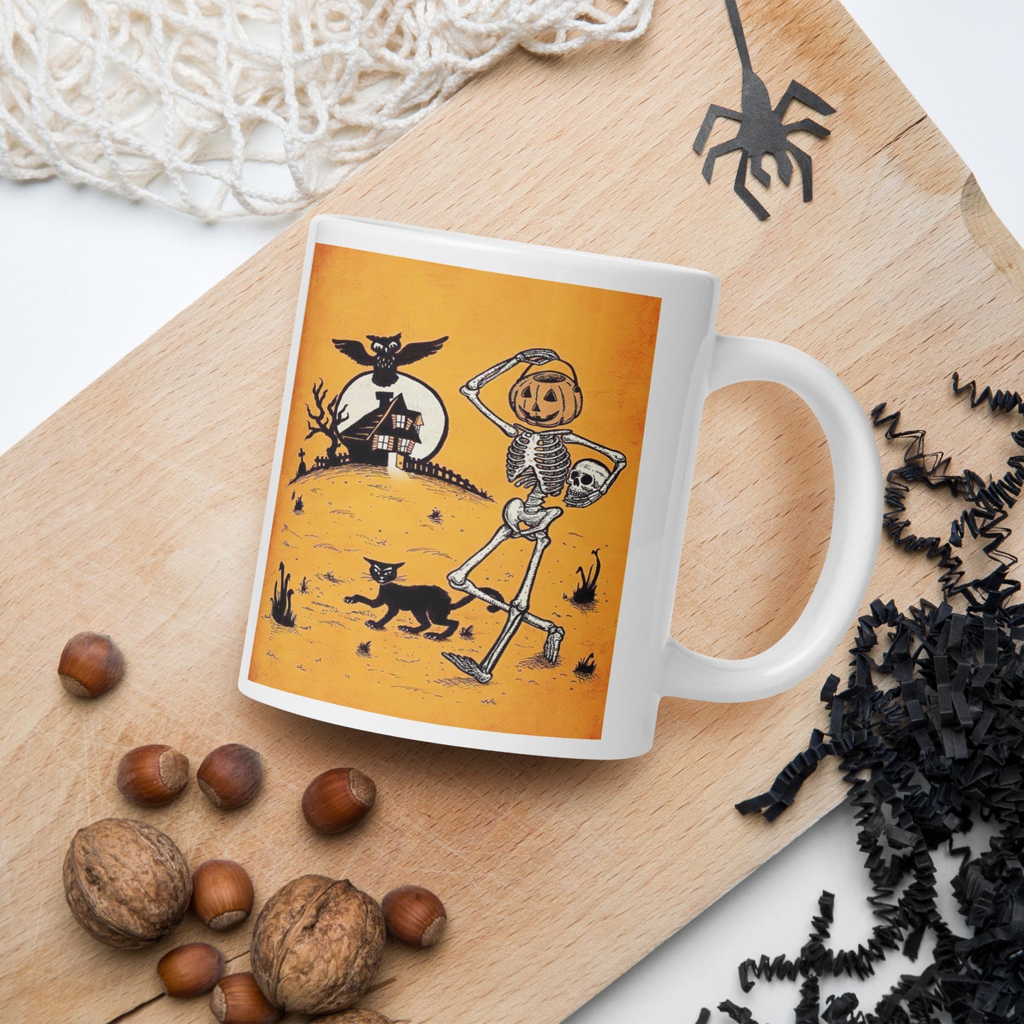 This ceramic glossy coffee mug features a vintage Halloween illustration of a skeleton with a jack o lantern for a head, a black cat, a black owl, a spooky house and a full moon. 