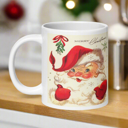 This ceramic glossy coffee mug features a midcentury retro Christmas illustration of Santa smiling under a sprig of mistletoe. It says Merry Christmas abovehis head and there are snowflakes.