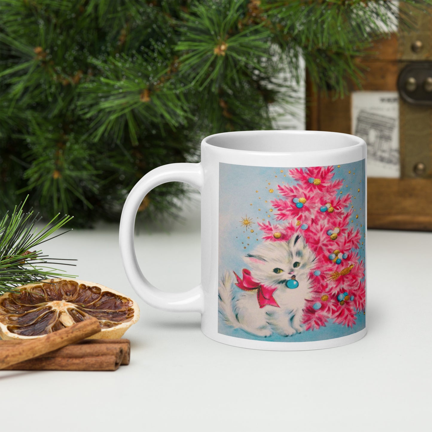 This glossy ceramic coffee cup features an adorable vintage retro print of a white christmas kitten with a blue ornament in her mouth. She is wearing a pink bow and is sitting next to a pink Christmas tree with a blue background.