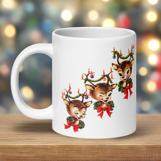 This ceramic glossy coffee mug features a vintage illustration of three adorable reindeer adorned with red bows and wreaths around their necks and garland, candles and ornaments in their antlers. The reindeer are staggered in a diagonal pattern.