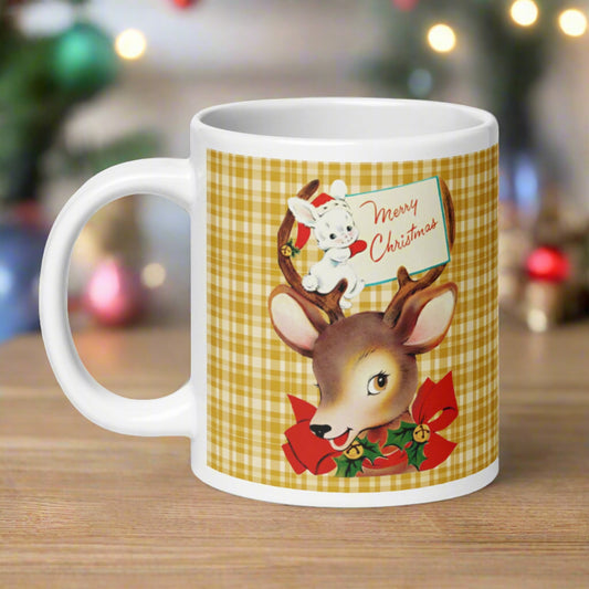 This ceramic glossy coffee mug features a retro vintage illustration of a reindeer wearing a red bow with bells and holly. There is a white bunny sitting in her antlers wearing a santa hat and mittens holding a card that says Merry Christmas.