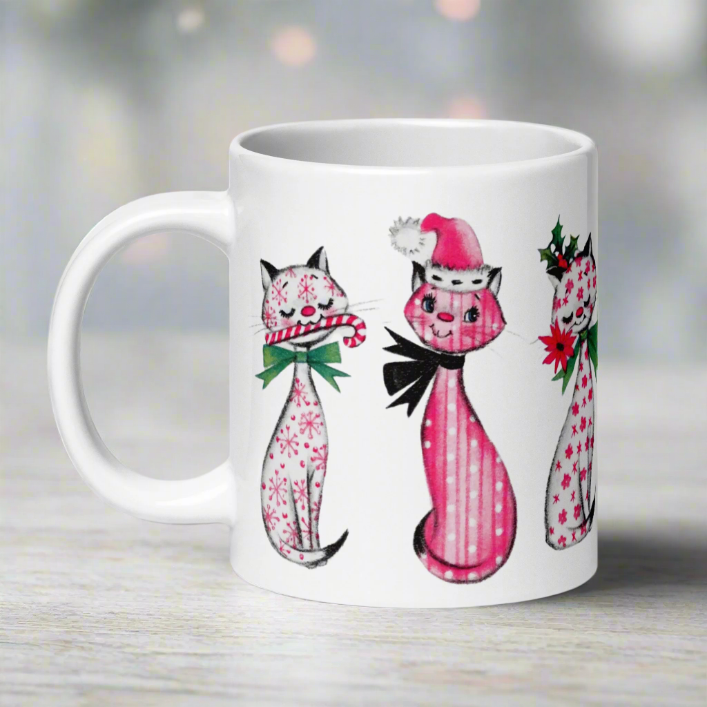 This ceramic glossy coffee mug features a midcentury retro Christmas illustration of three cats. They are adorned with bows around their necks. One has a pink santa hat on her head, one has a candy cane in her mouth and the other is holding a flower.