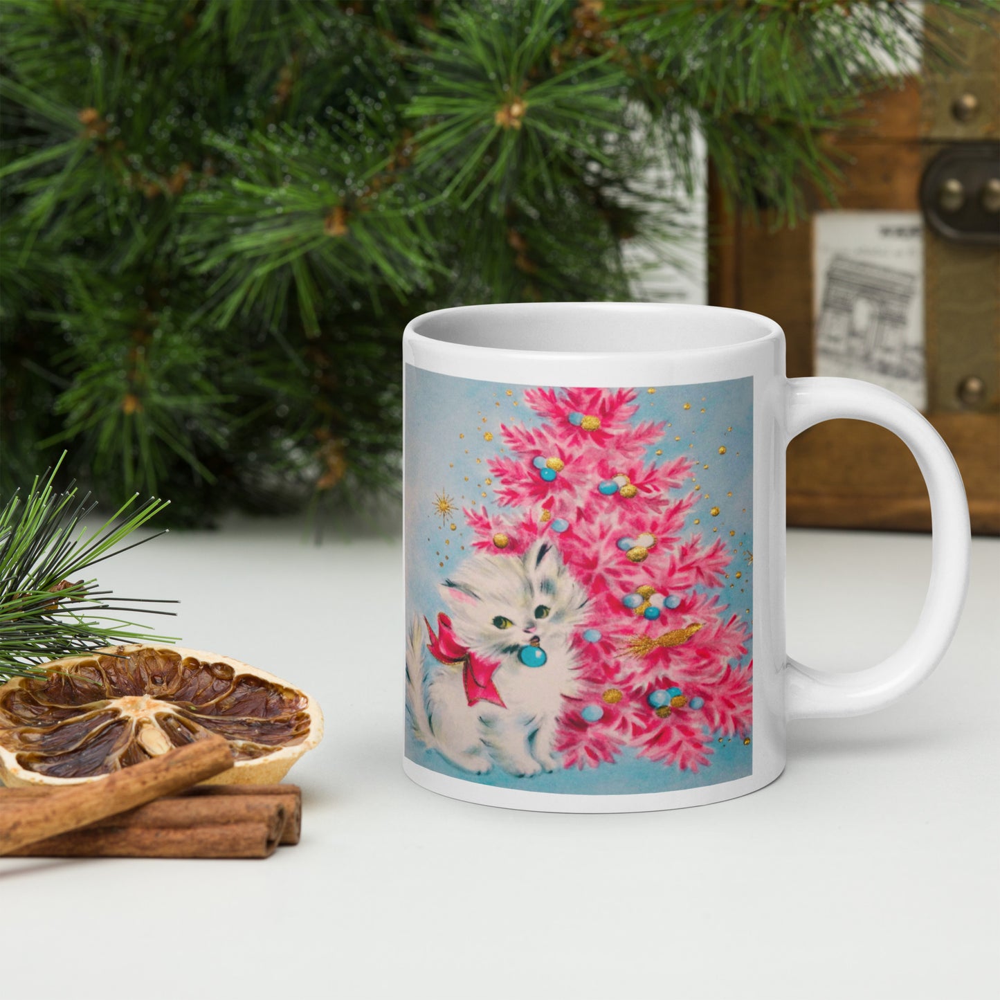 This glossy ceramic coffee cup features an adorable vintage retro print of a white christmas kitten with a blue ornament in her mouth. She is wearing a pink bow and is sitting next to a pink Christmas tree with a blue background.