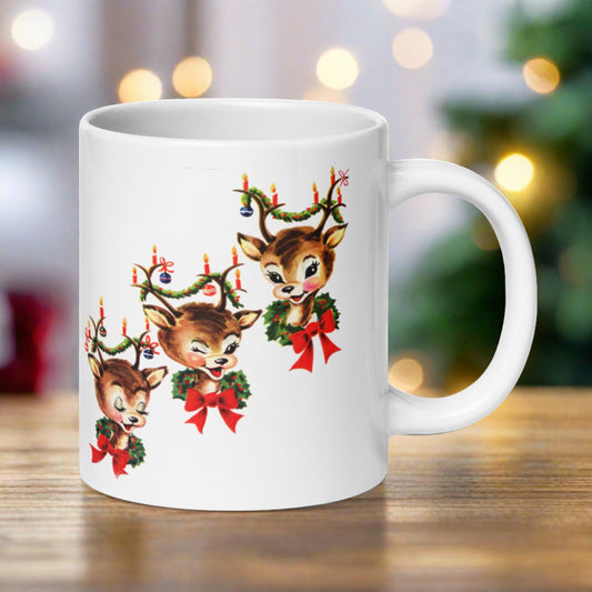 This ceramic glossy coffee mug features a vintage illustration of three adorable reindeer adorned with red bows and wreaths around their necks and garland, candles and ornaments in their antlers. The reindeer are staggered in a diagonal pattern.